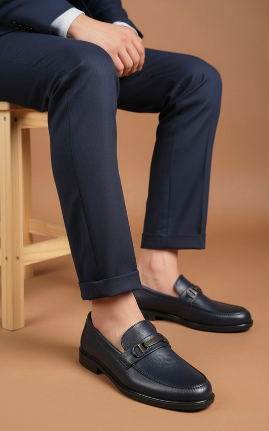 Person wearing navy blue dress shoes and pants sitting on a wooden stool against a brown background