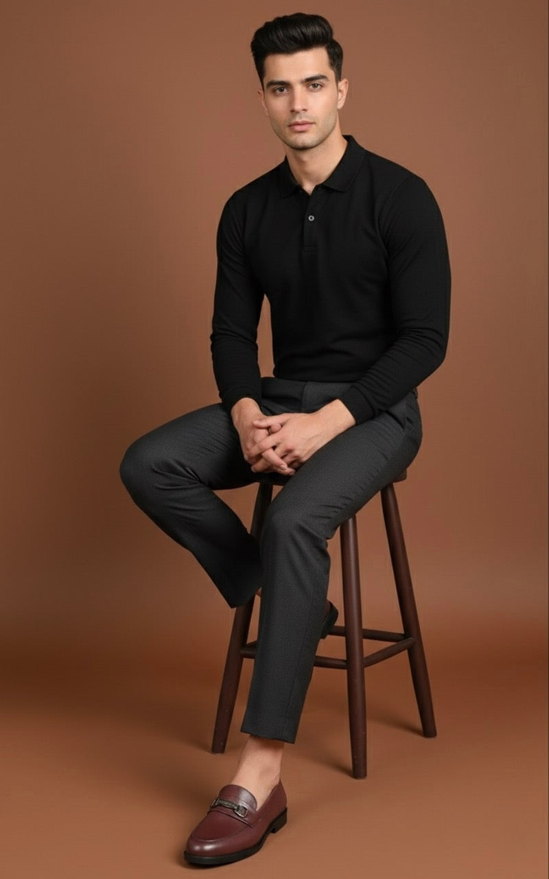 Man sitting on a stool wearing a black long-sleeve shirt and dark pants against a brown background