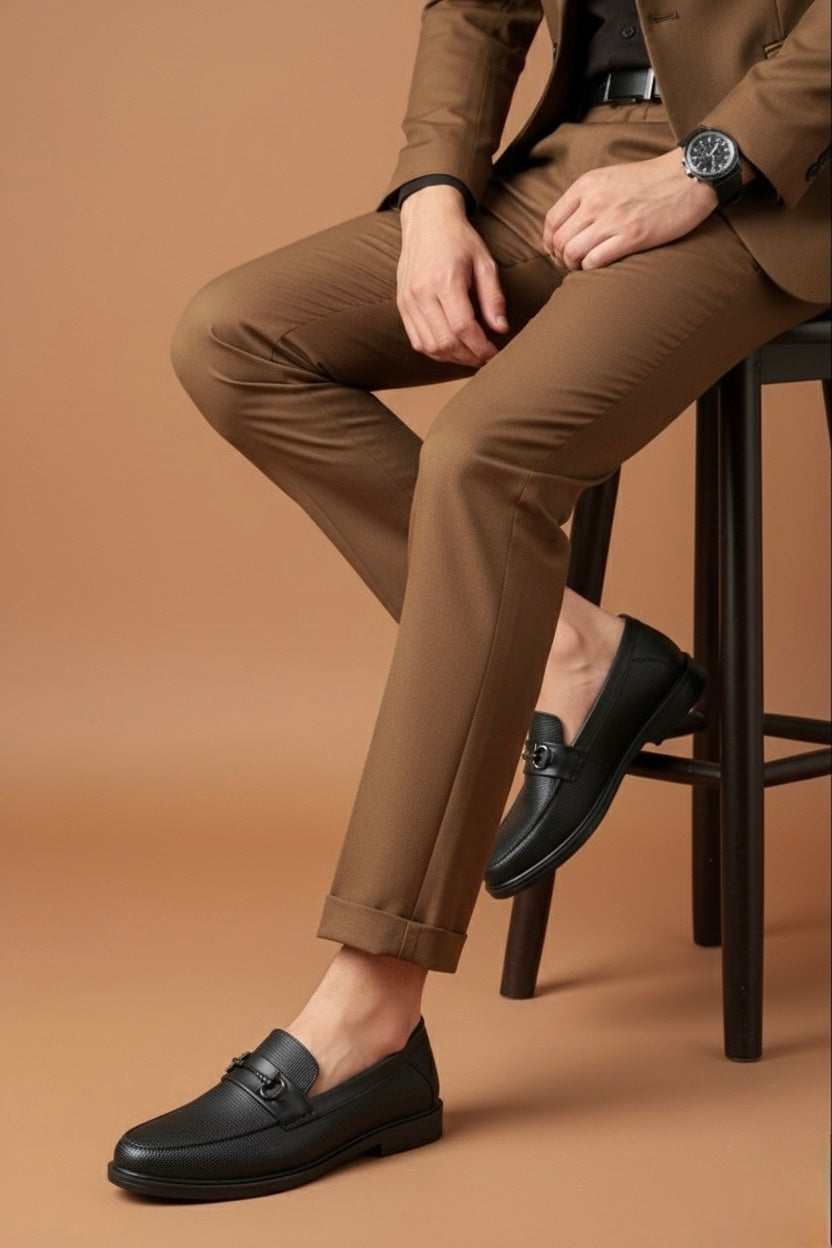 Person wearing a brown suit and black loafers sitting on a stool against a brown background