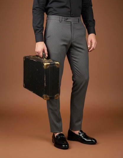 Person wearing gray trousers and black shoes holding a vintage suitcase on a brown background