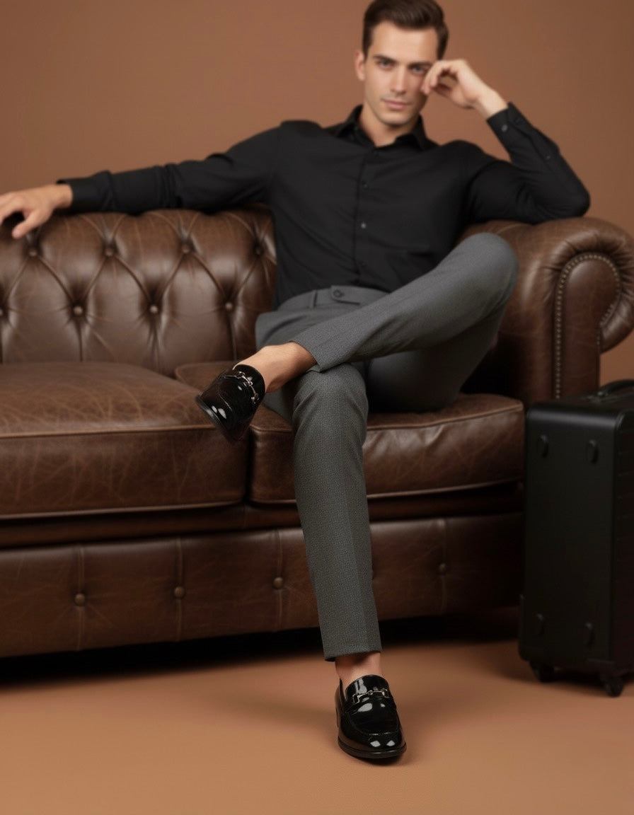 Man in black shirt and gray pants sitting on a brown leather couch with a suitcase next to him.
