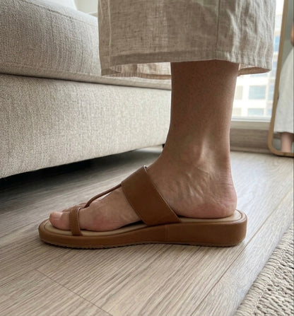 Person wearing a brown flat kolhapuri sliders sandal on a wooden floor with a beige sofa in the background