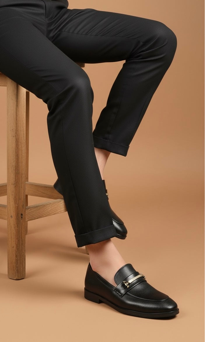 Person wearing black dress shoes and pants sitting on a wooden stool against a beige background