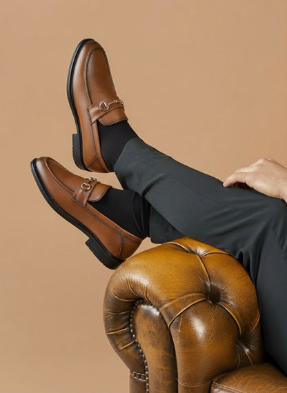 Brown leather shoes with a gold buckle worn by a person sitting on a brown leather ottoman against a beige background.