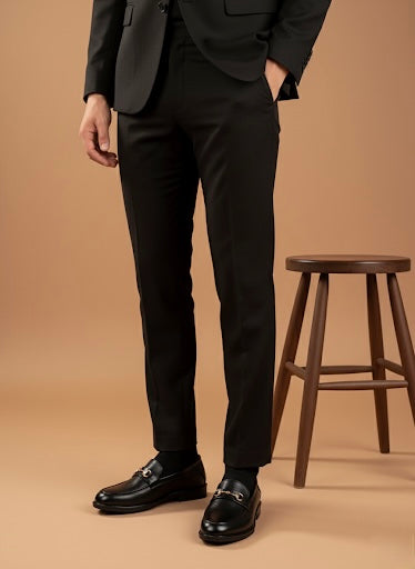 Person wearing a black suit and shoes standing next to a wooden stool on a beige background