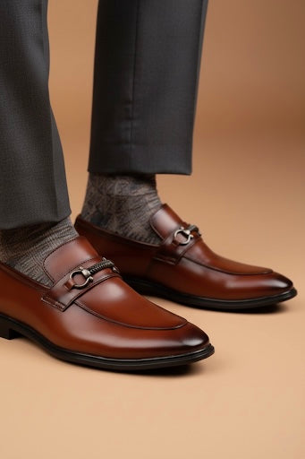 Brown dress shoes with silver buckles worn with dark gray pants and socks on a beige background