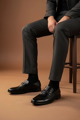 Person wearing black dress shoes and gray pants sitting on a brown stool against a brown background