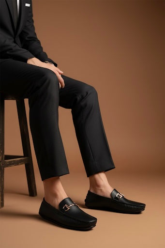 Person wearing a black suit and loafers sitting on a stool against a brown background