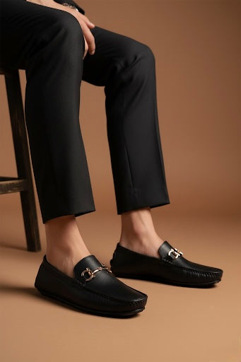 Person wearing black loafers with a gold buckle on a brown background
