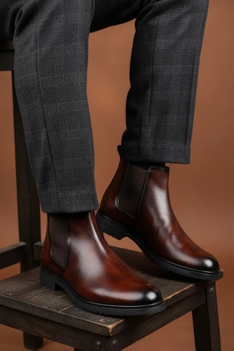 Brown leather Chelsea boots worn with gray checkered pants on a wooden chair against a brown background